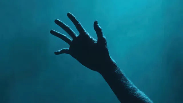 Silhouetted hand reaching upward into dim blue light with visible skin texture against a dark background.