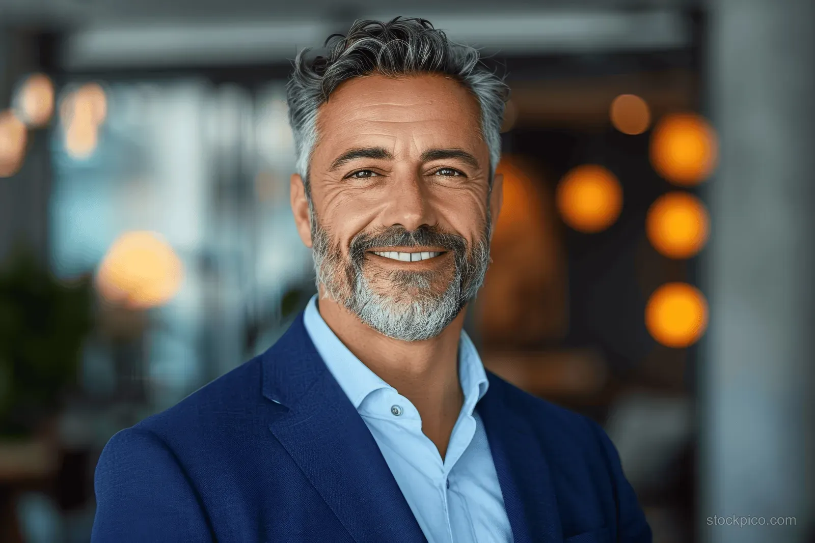 Confident middle-aged businessman with gray hair and a goatee smiles in a blue suit against a softly blurred modern office background.