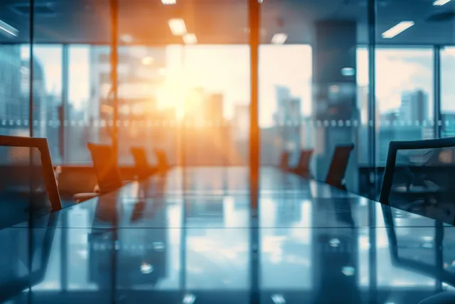 Defocused conference room chairs and tables beside glass walls with sunlight and bokeh from office windows.