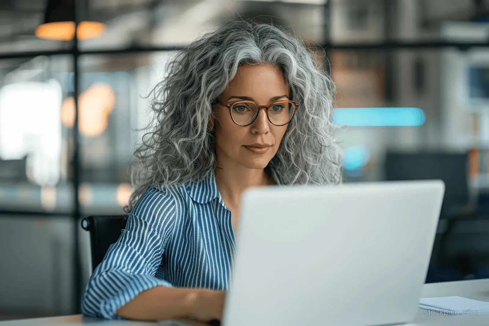 Grey-haired businesswoman with glasses types on a laptop at a desk in a modern office.