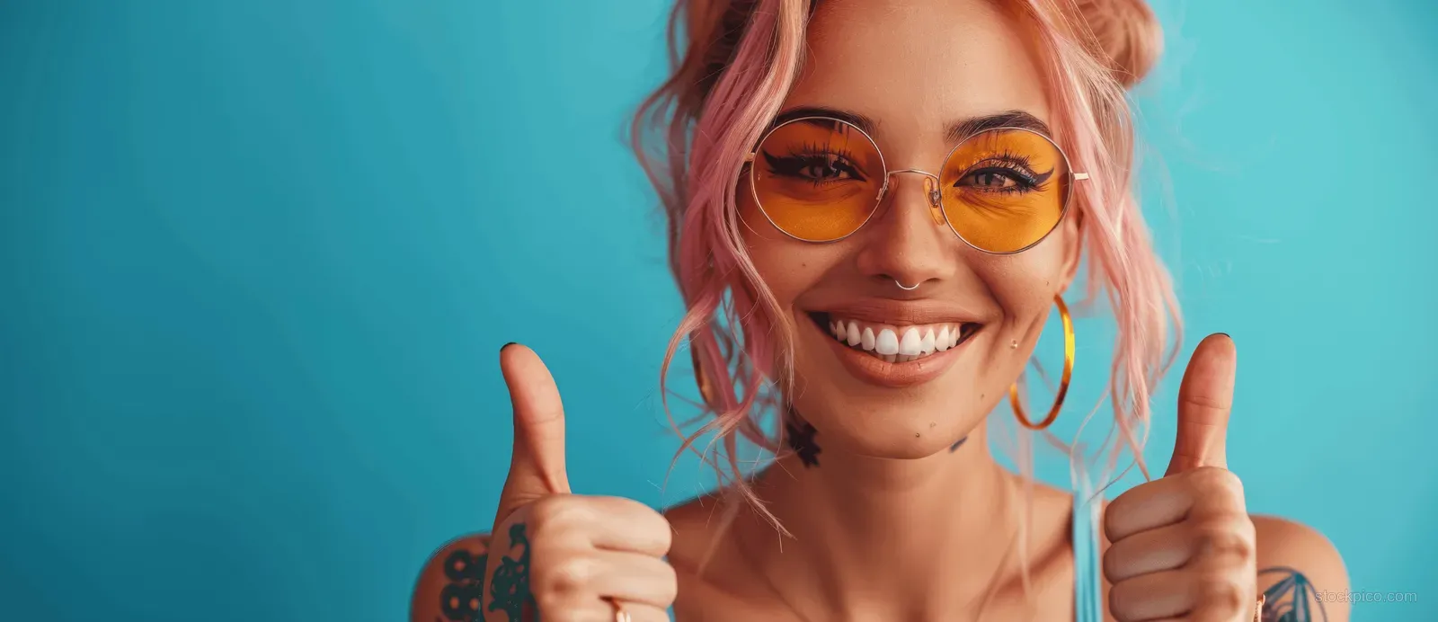Smiling punk woman with pink hair and sunglasses gives double thumbs up against a blue background.