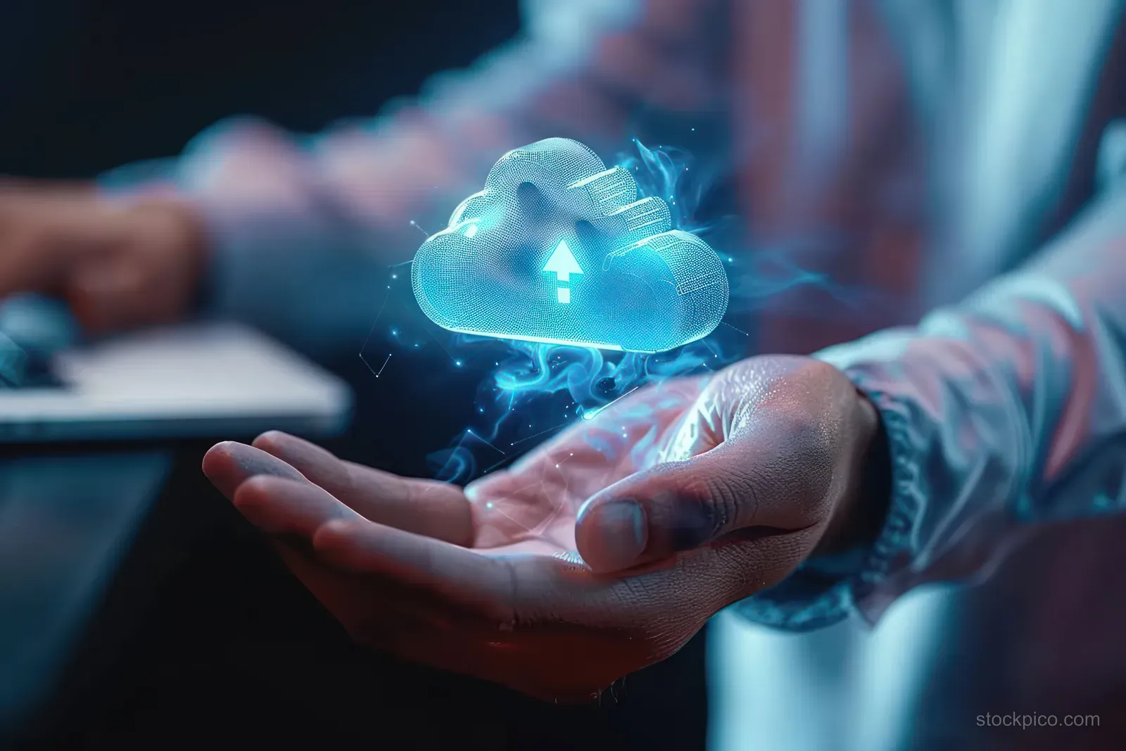 A person’s hand holds a glowing cloud icon with an upload arrow over a laptop workspace in the background.