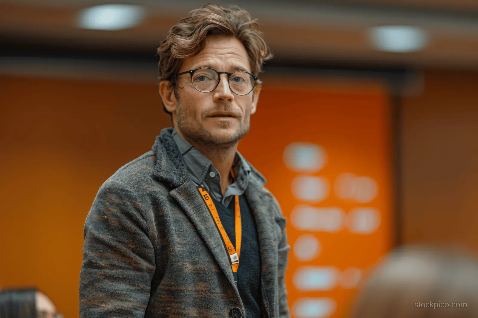 A man with glasses and wavy hair stands indoors wearing a gray sweater and orange lanyard, looking at the camera.