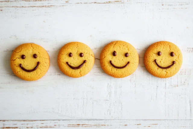 Four smiley face cookies with chocolate eyes and smiles arranged in a row on a rustic wooden table.