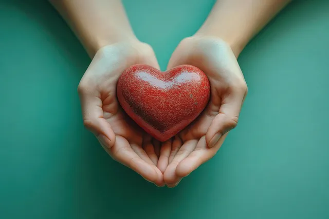 Two hands hold a red heart icon against a teal background symbolizing care and donation.