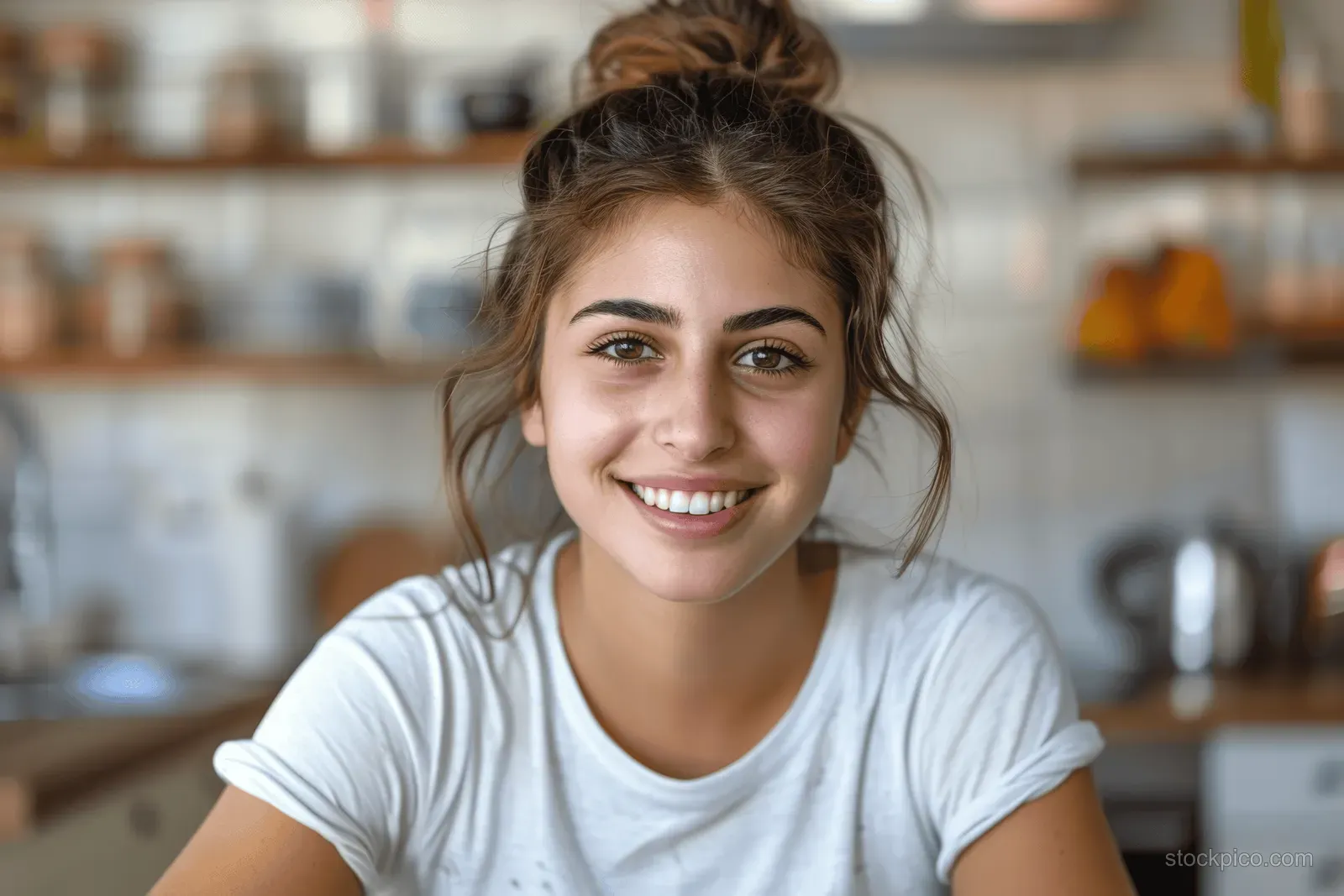 A smiling young woman with brown hair in a bun relaxes in a cozy, well-lit kitchen.