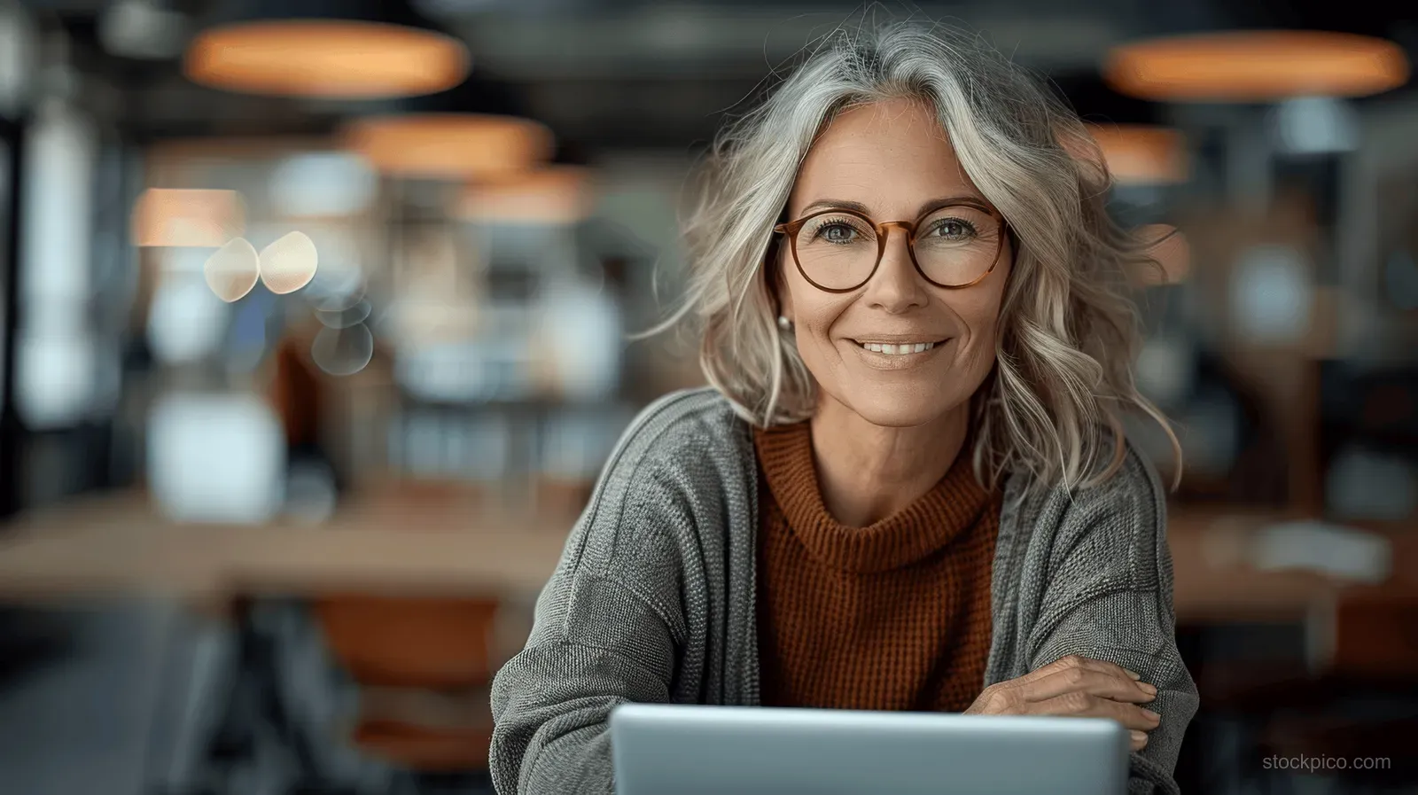 Smiling senior businesswoman holding a tablet while seated at a desk in a softly lit office.
