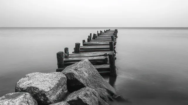 Weathered wooden pier extends into calm ocean, framed by large rocks in monochrome