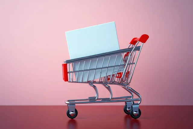 A small metallic shopping cart with red accents holds a blank white rectangular box against a pink background.