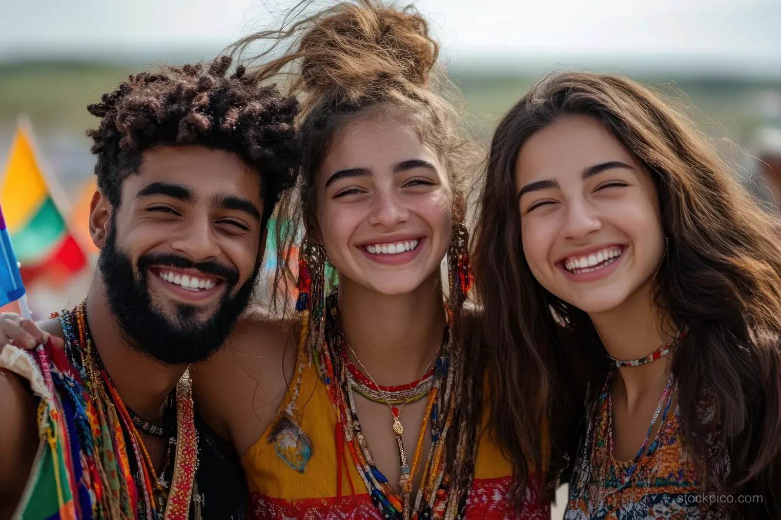 Three young adults smile together outdoors while wearing colorful bohemian clothing and jewelry with rainbow flags behind them.