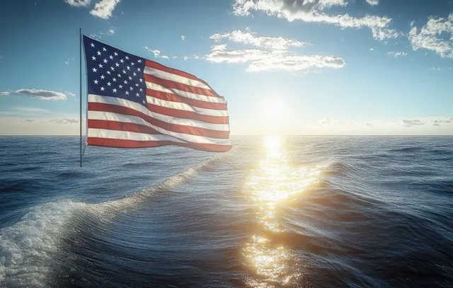 An American flag billows above ocean waves with sunlight reflecting across the rippling water under a partly cloudy sky.