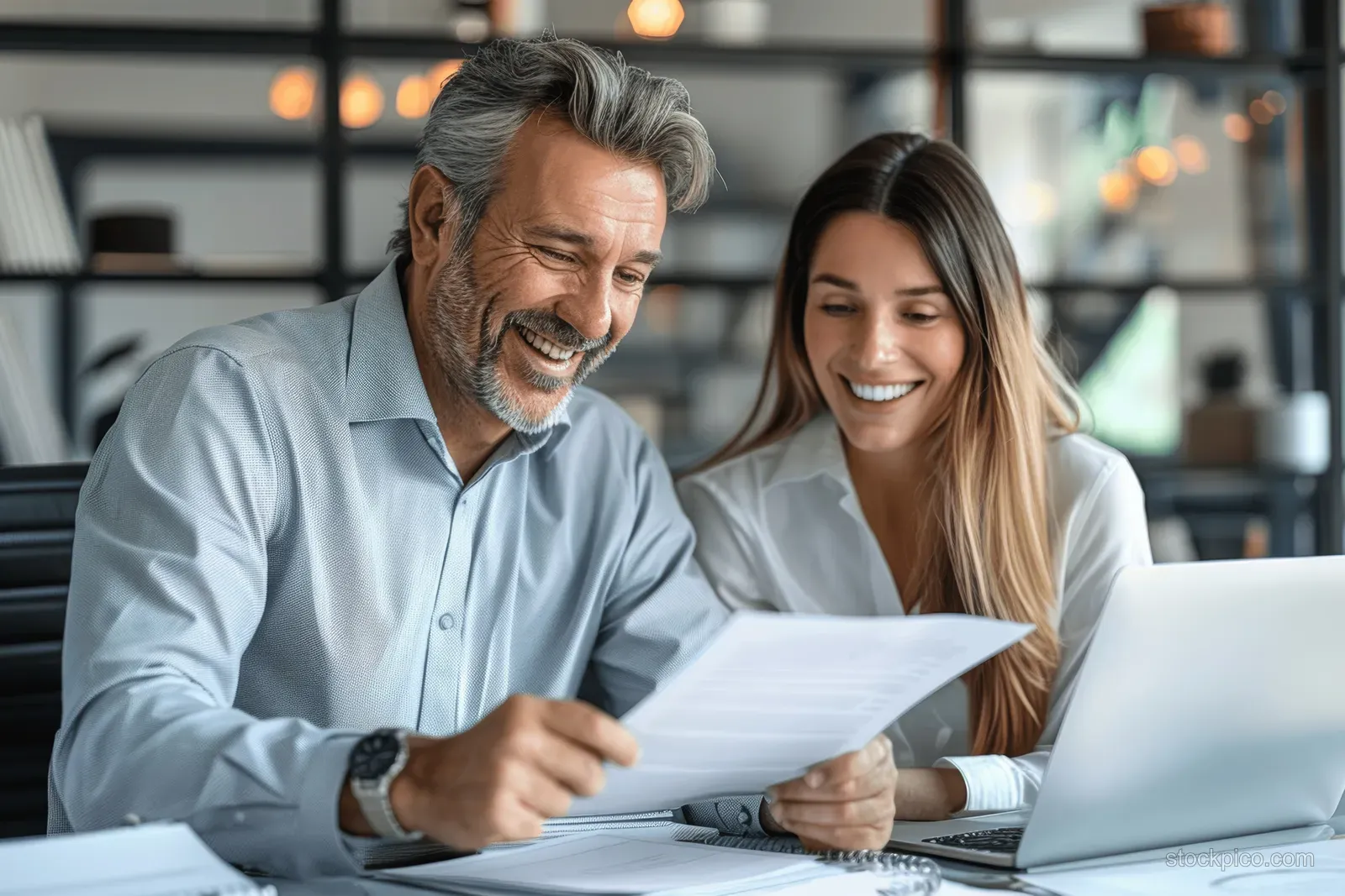 A smiling manager and coworker review printed financial documents at an office desk beside a laptop.
