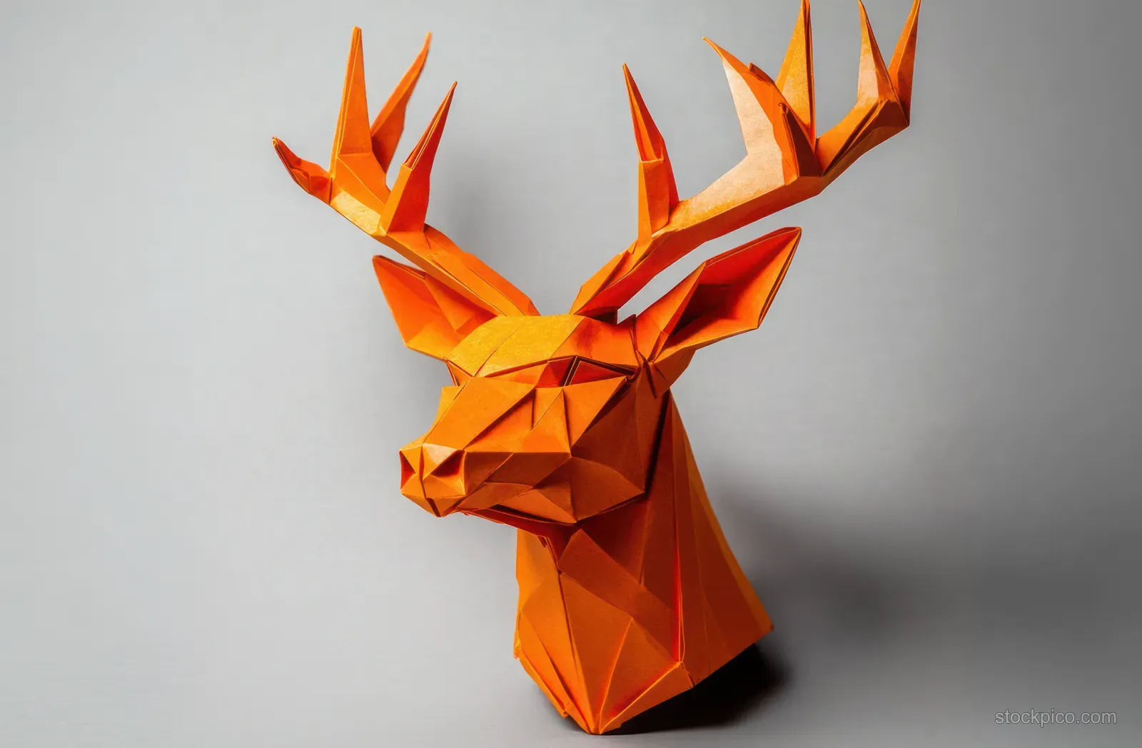 Orange geometric paper sculpture of a deer head with large antlers on a plain light gray background.