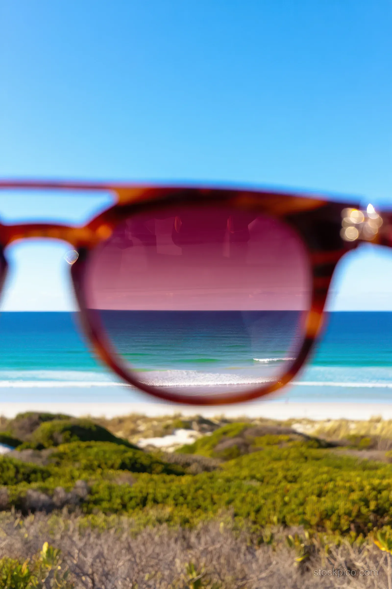 Sunglasses held up with the ocean and beach visible through tinted lenses under a clear blue sky.