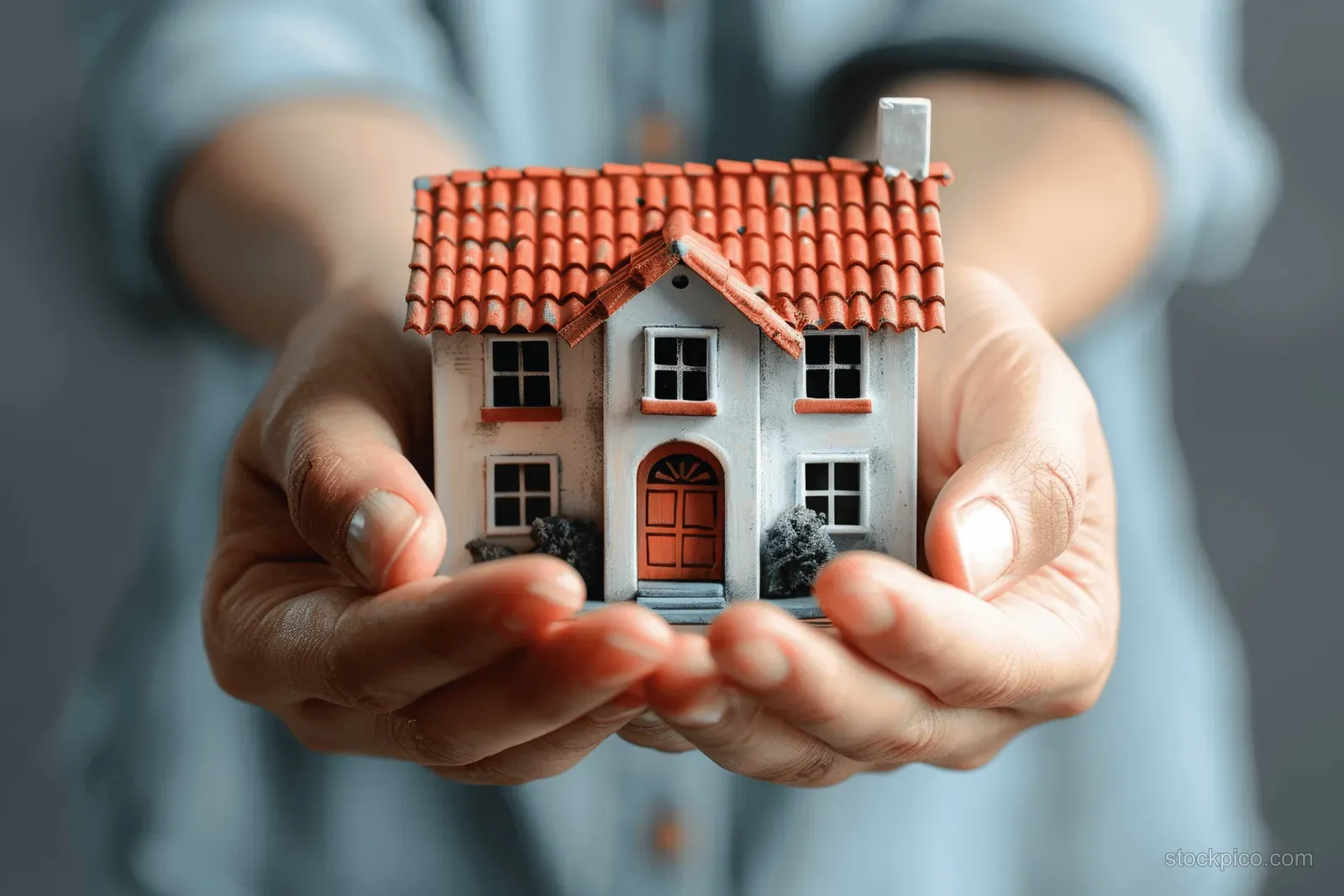 Person’s hands cradle a miniature house model with a red roof against a plain gray background.