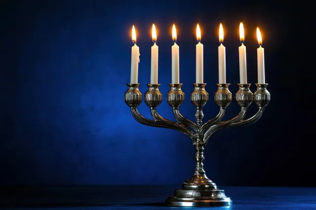 A silver menorah with seven lit white candles glows against a dark blue background.