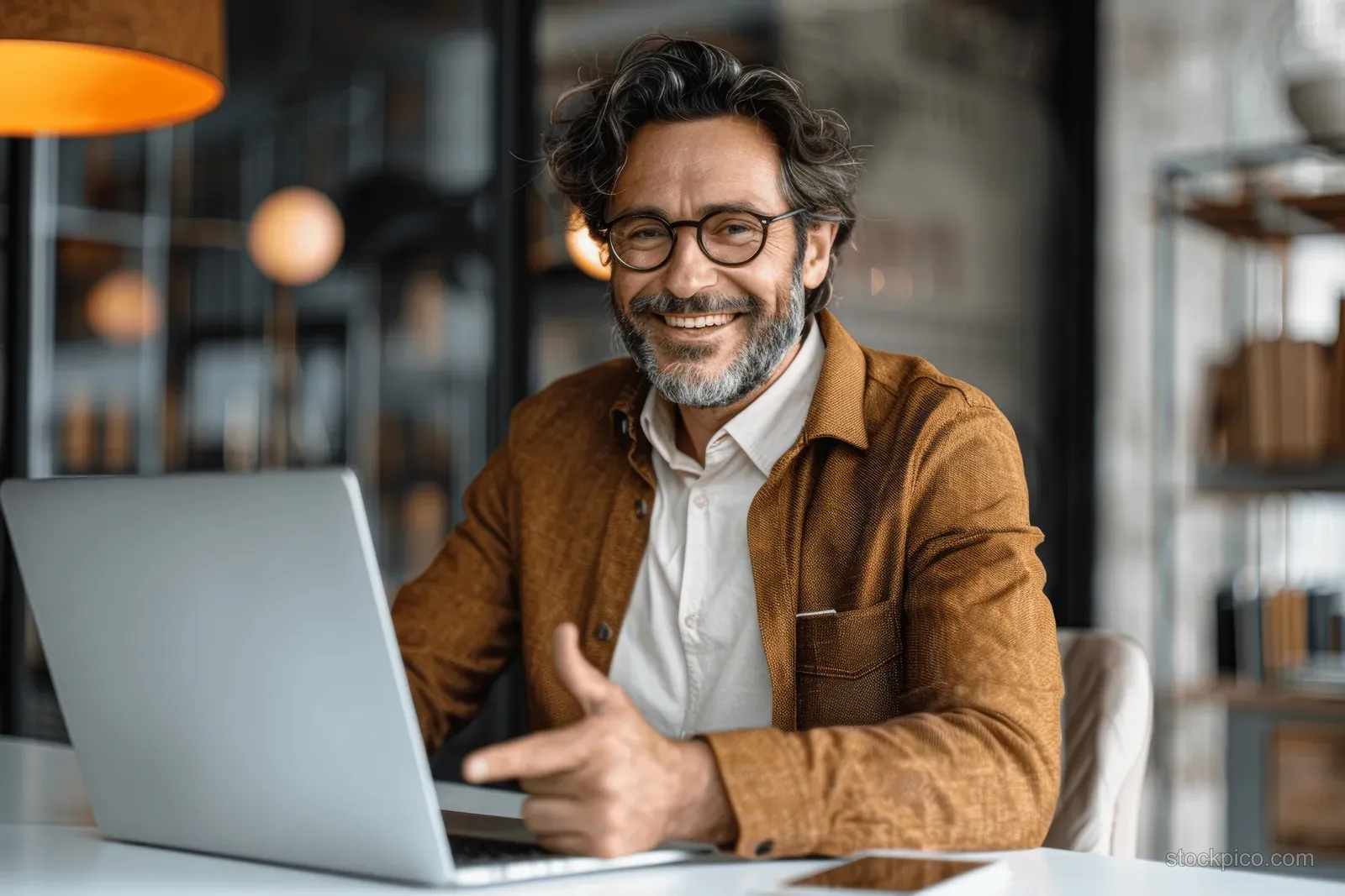 A smiling businessman in glasses gestures beside a laptop during a video call in an office.