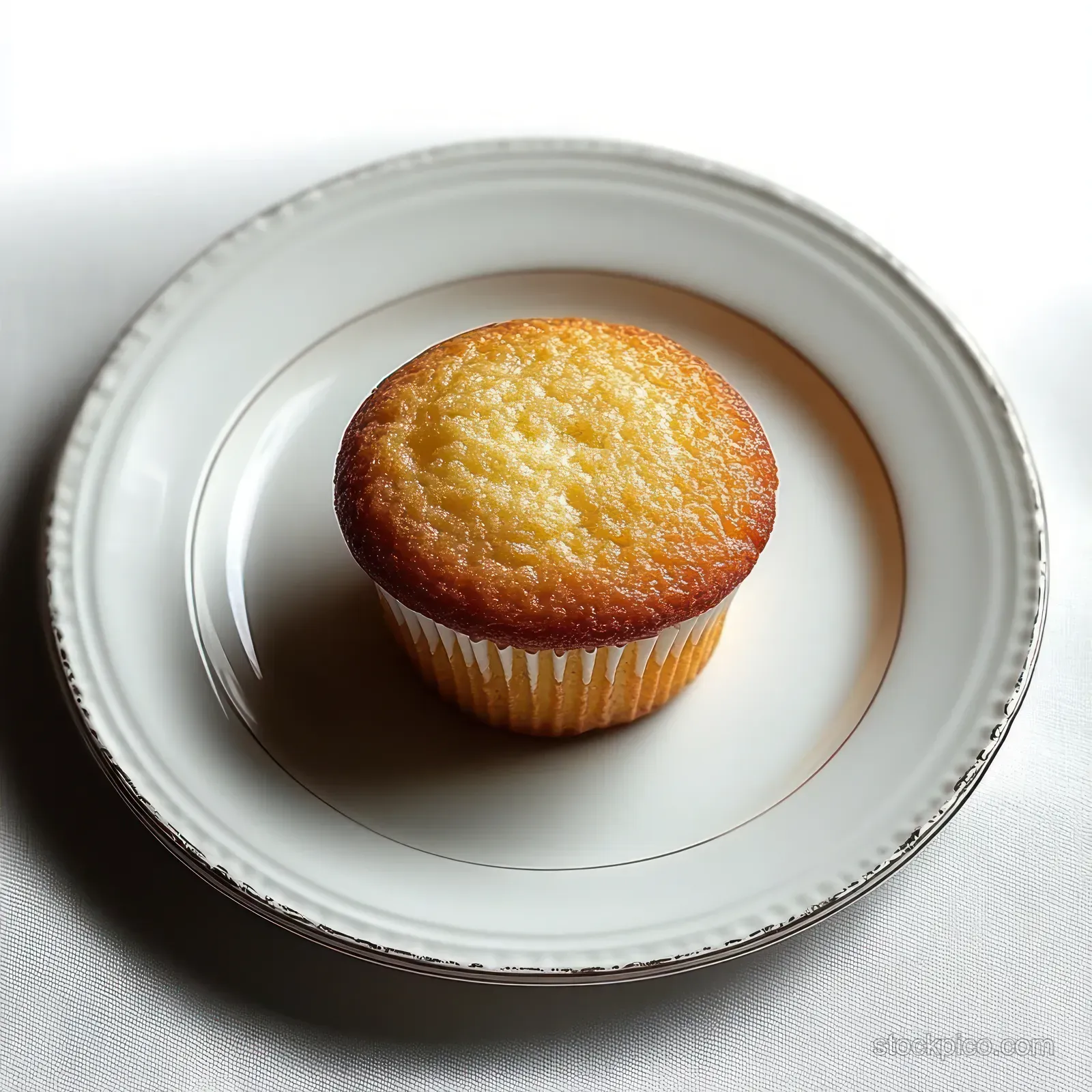 A golden-brown cupcake with a crisp top on a white porcelain plate under soft light.