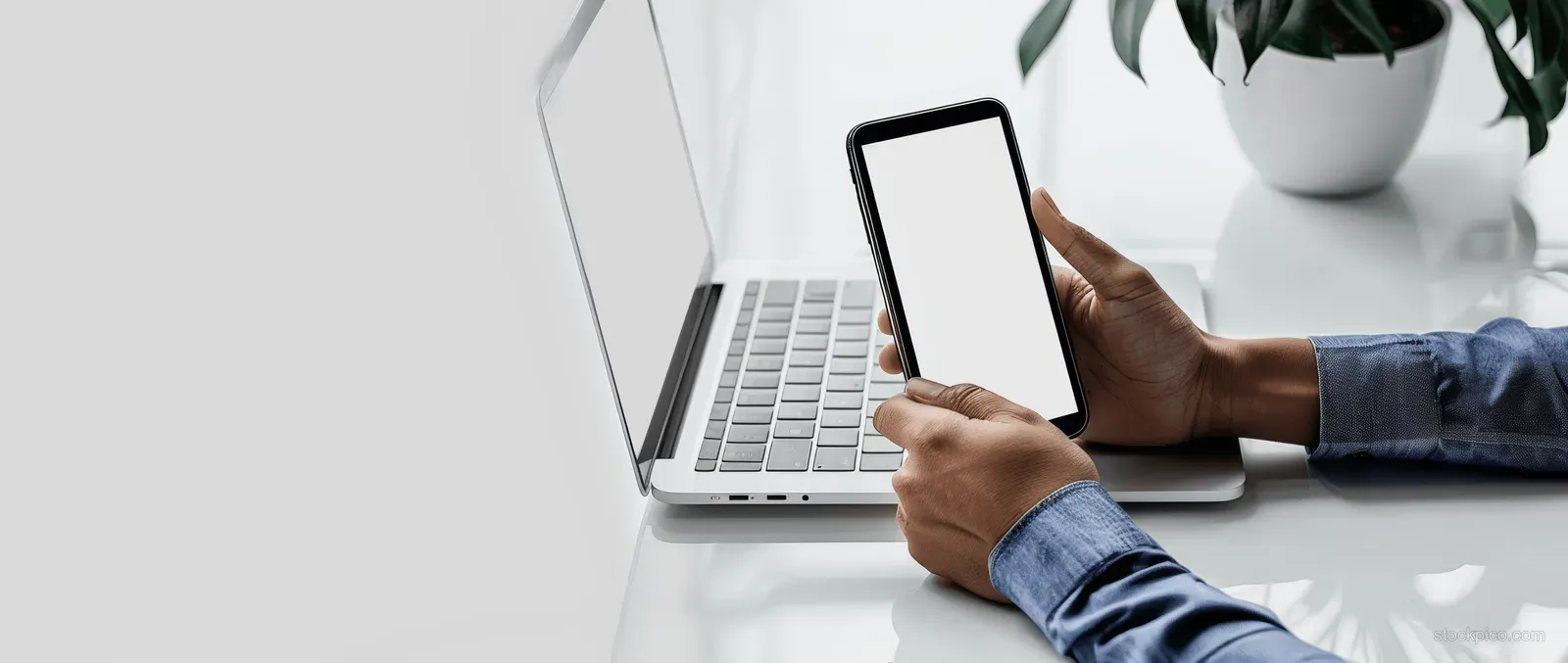 A person types on a laptop while holding a smartphone at a desk in an office.