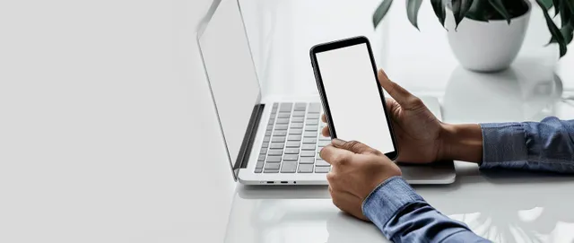 A person types on a laptop while holding a smartphone at a desk in an office.