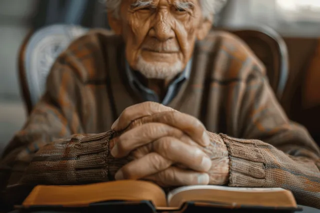 Elderly man kneeling with hands clasped over an open Holy Bible on a table at home.