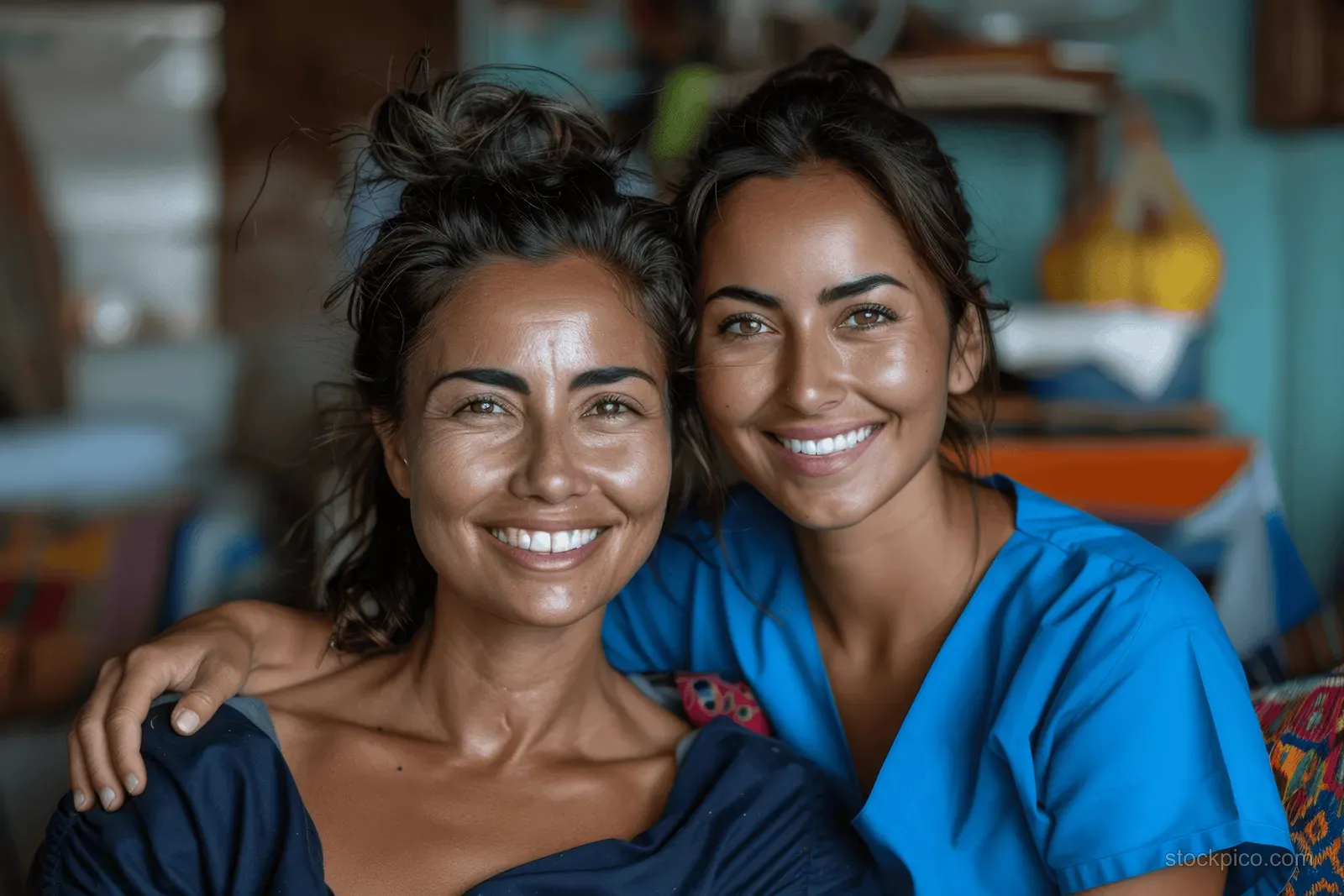 Nurse in blue scrubs hugs an elderly woman while both smile indoors in a home care setting.
