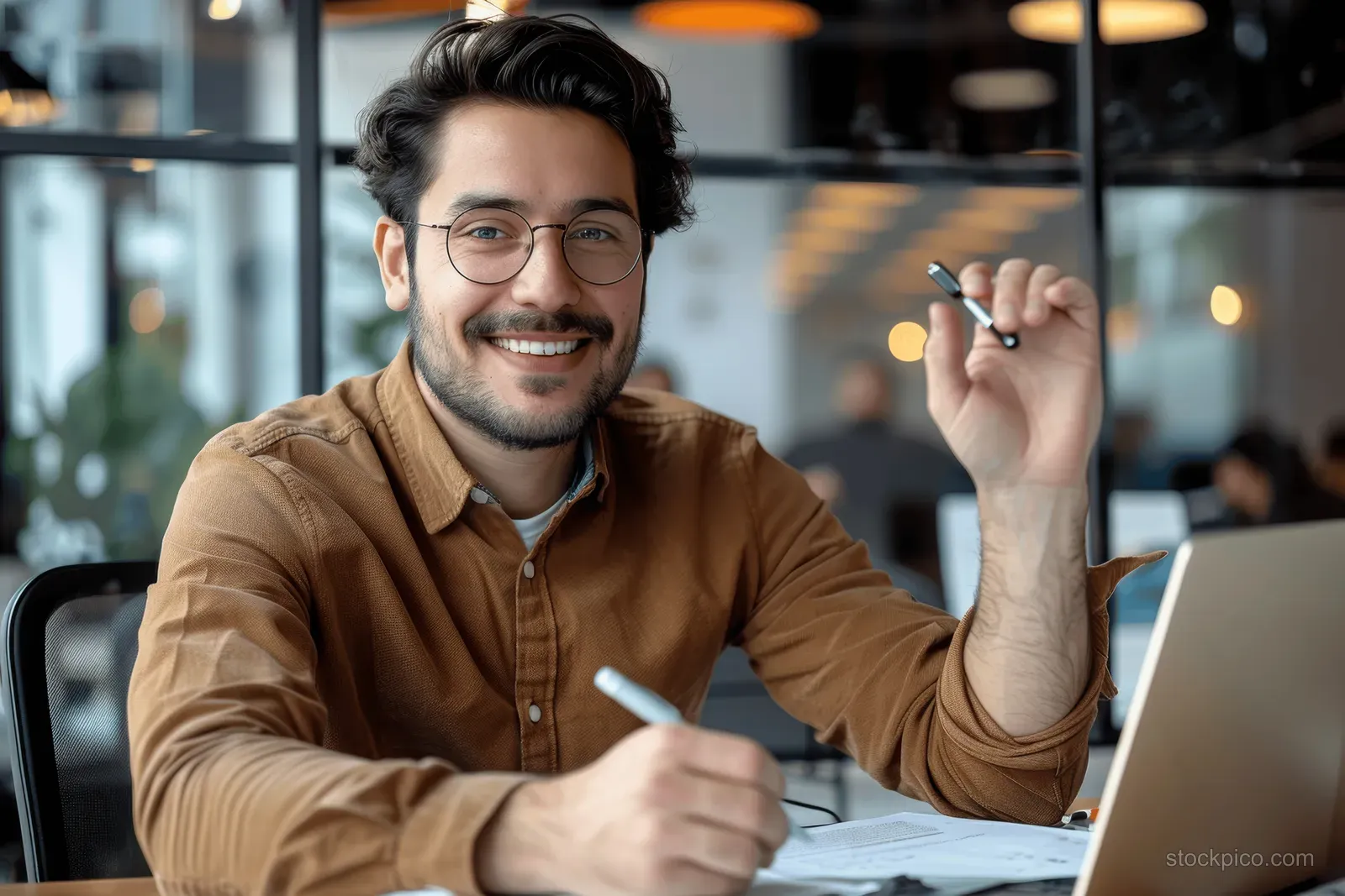 Smiling young man at a desk using a laptop while reviewing paperwork in a modern office with window light.