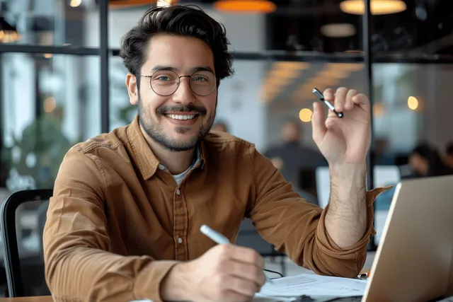 Smiling young professional at laptop desk, reviewing paperwork with calculator in bright office