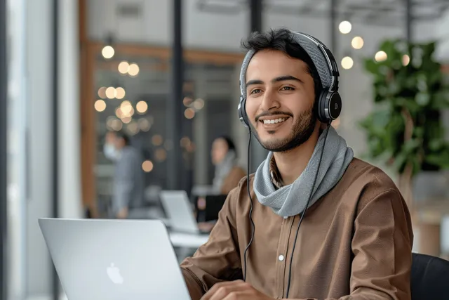 Cheerful young Arab man wearing a headset sits at a desk using a laptop during an online video call.
