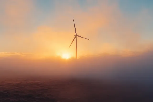 A lone wind turbine emerges from sunrise fog over a misty landscape with orange and blue sky.