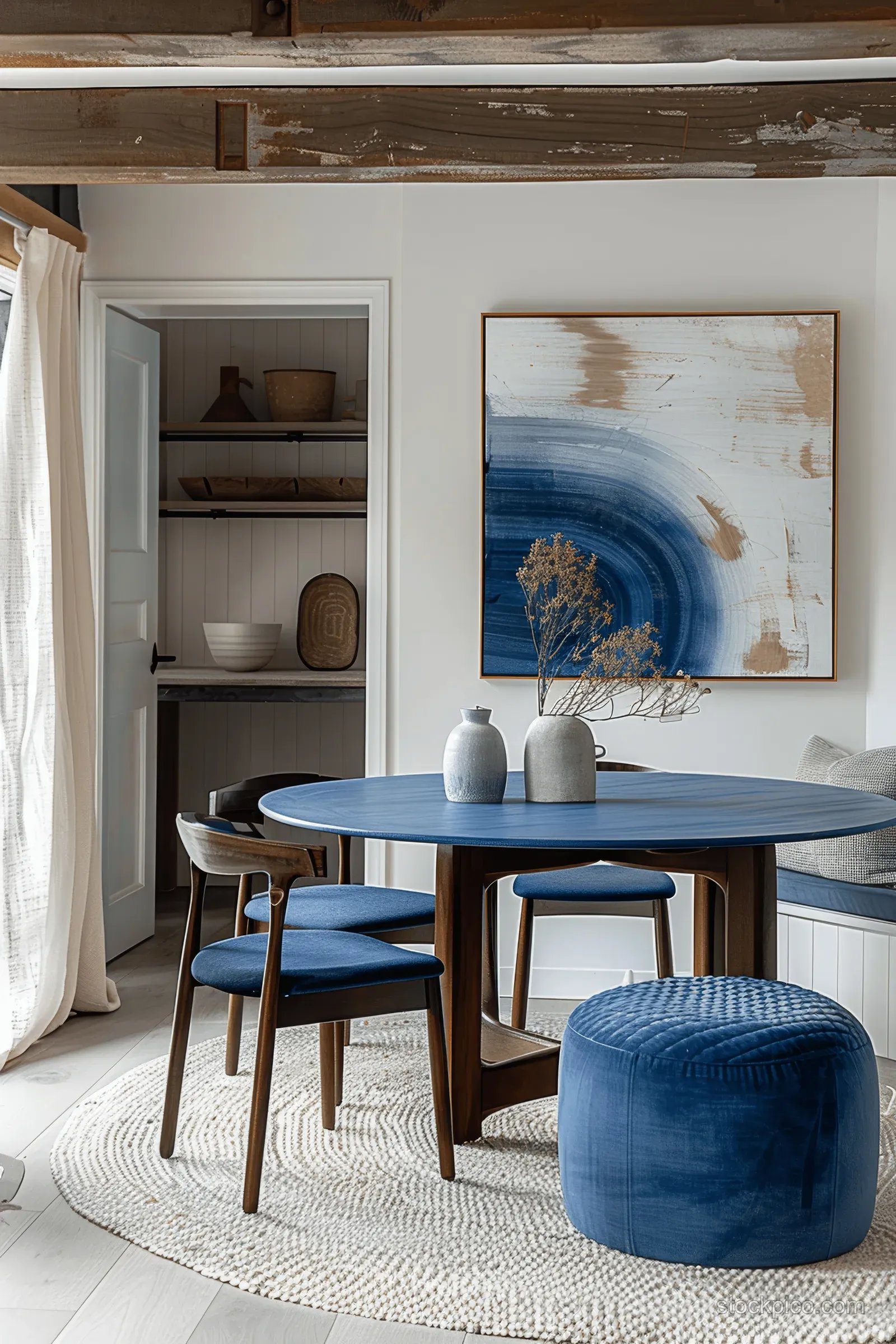Blue dining table with wooden chairs and a round ottoman beside a white wall with abstract painting.