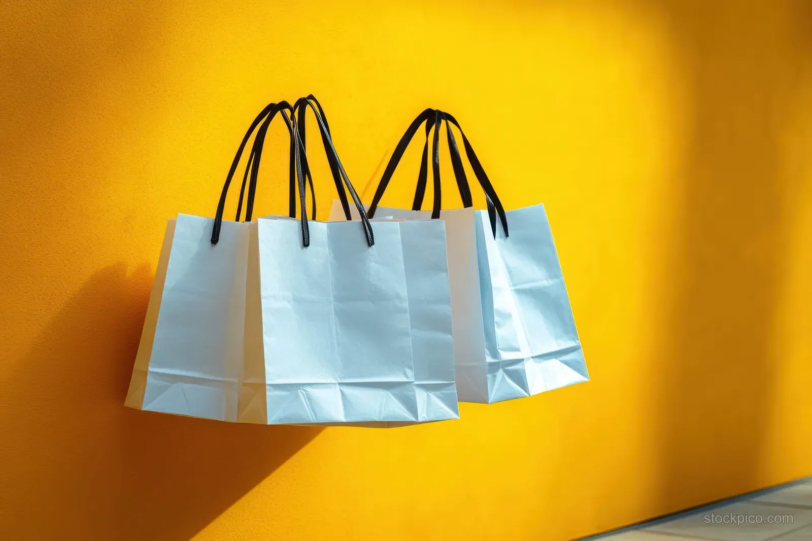 Three white paper shopping bags with black handles hang against a bright yellow wall with soft shadows.