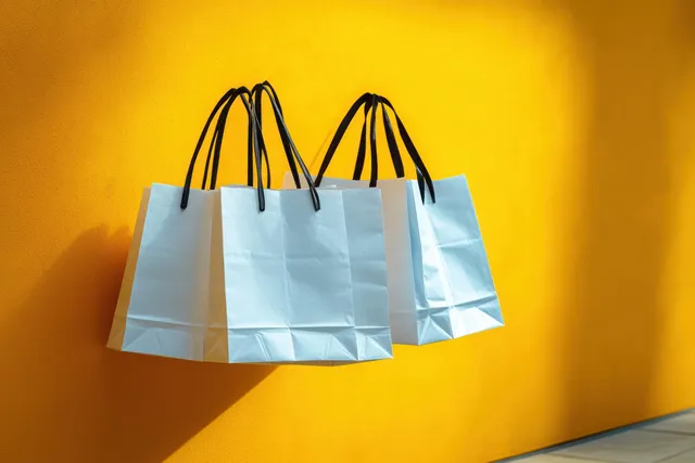 Three white paper shopping bags with black handles hang against a bright yellow wall with soft shadows.