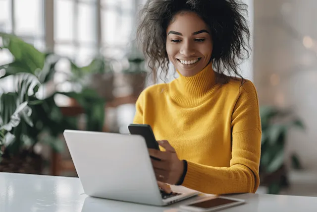 Young woman typing on laptop while using smartphone at desk, blurred office background
