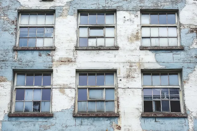 A decaying building facade with six multi-pane windows, peeling blue paint, and rusty window frames.