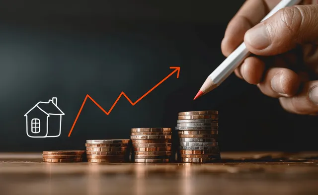A hand draws an upward growth graph beside a house icon and stacked coins on a desk.