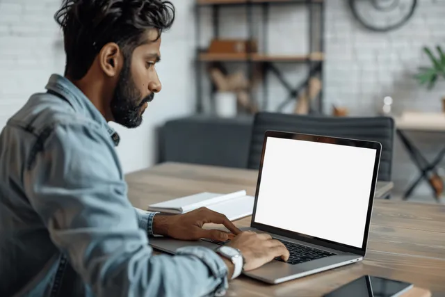 An Indian man works on a laptop with a blank screen while taking notes at a desk in a modern office.