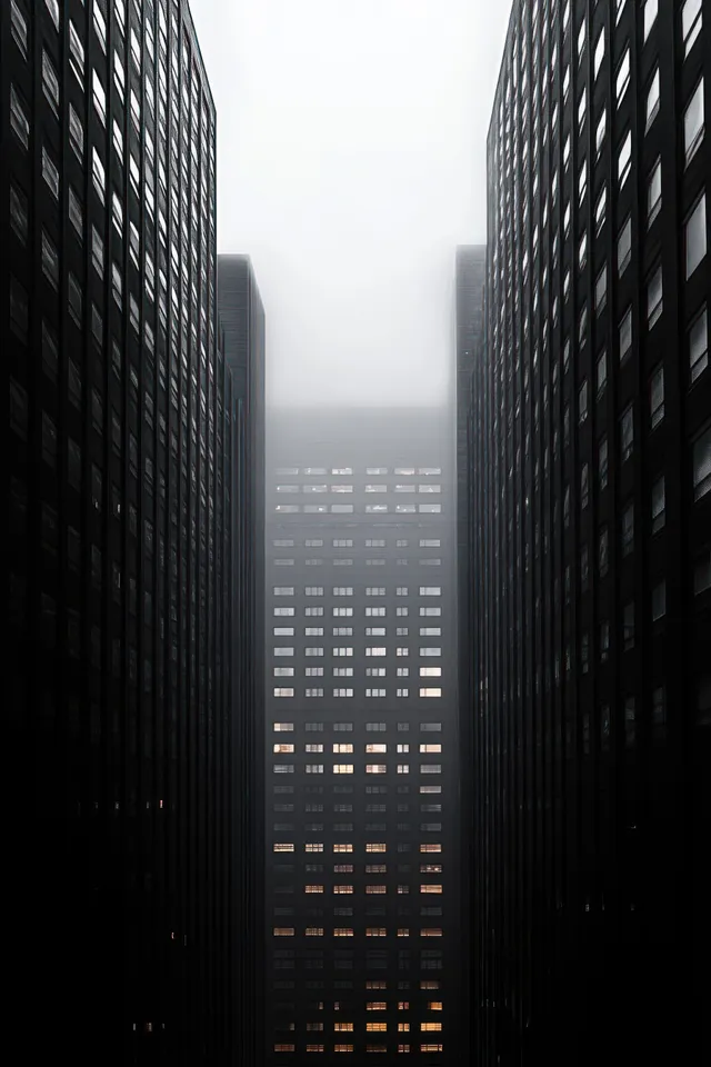 Foggy skyline view of tall buildings with glowing windows visible between two dark high-rises.