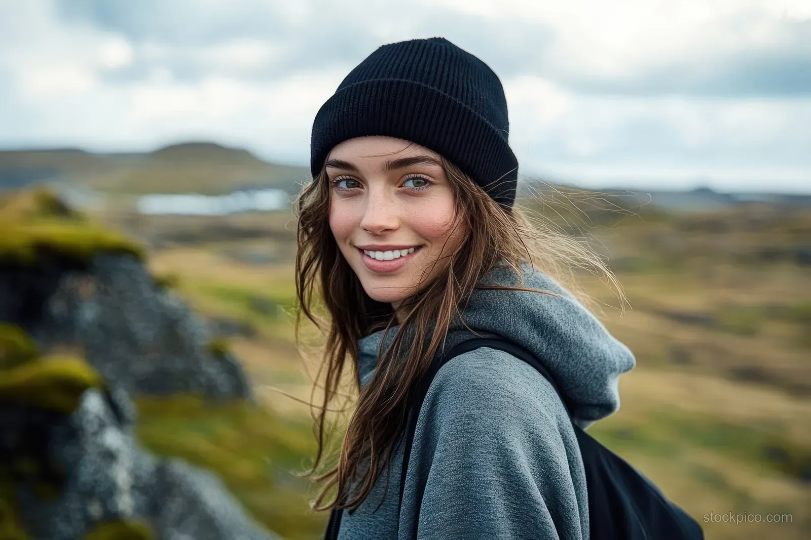 Smiling young woman in a black beanie and gray hoodie stands outdoors with cloudy hills and grassy landscape behind her.