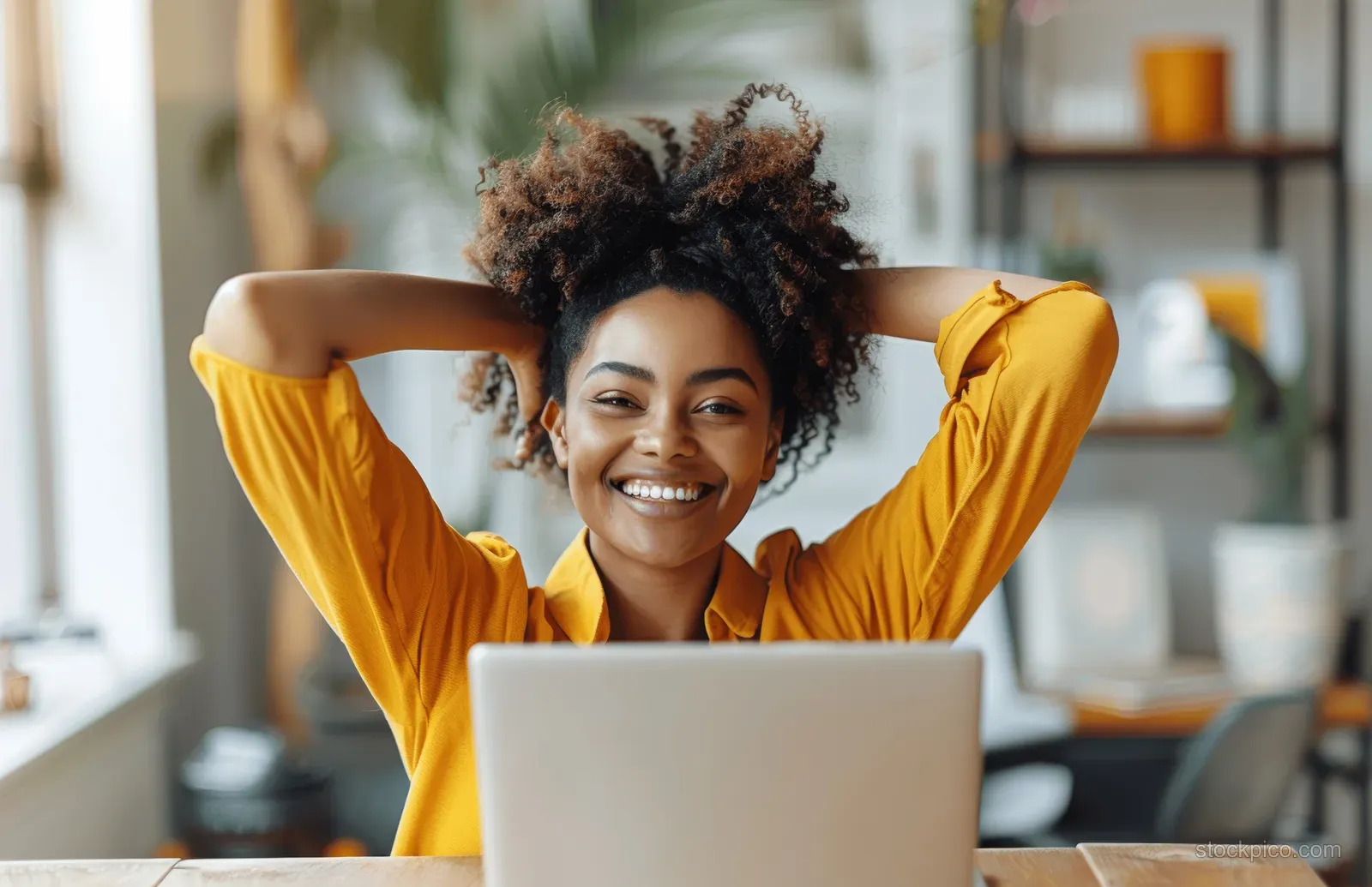 A smiling African American woman celebrates with arms raised behind a laptop in her home office.