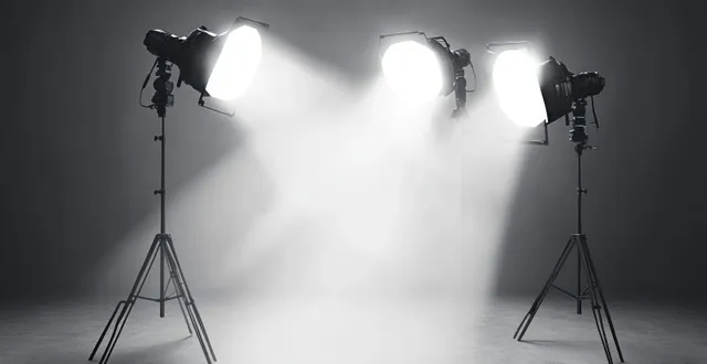 Three studio spotlights on tripods beam bright white light through a dark empty room.
