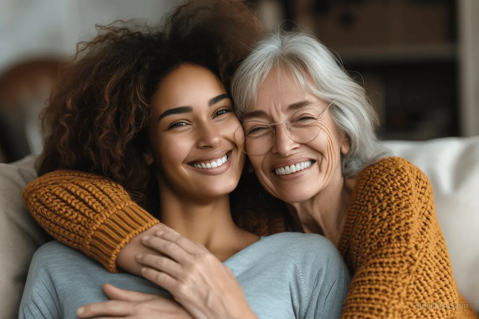 Senior mother and adult daughter embrace and laugh on a couch in a cozy living room.