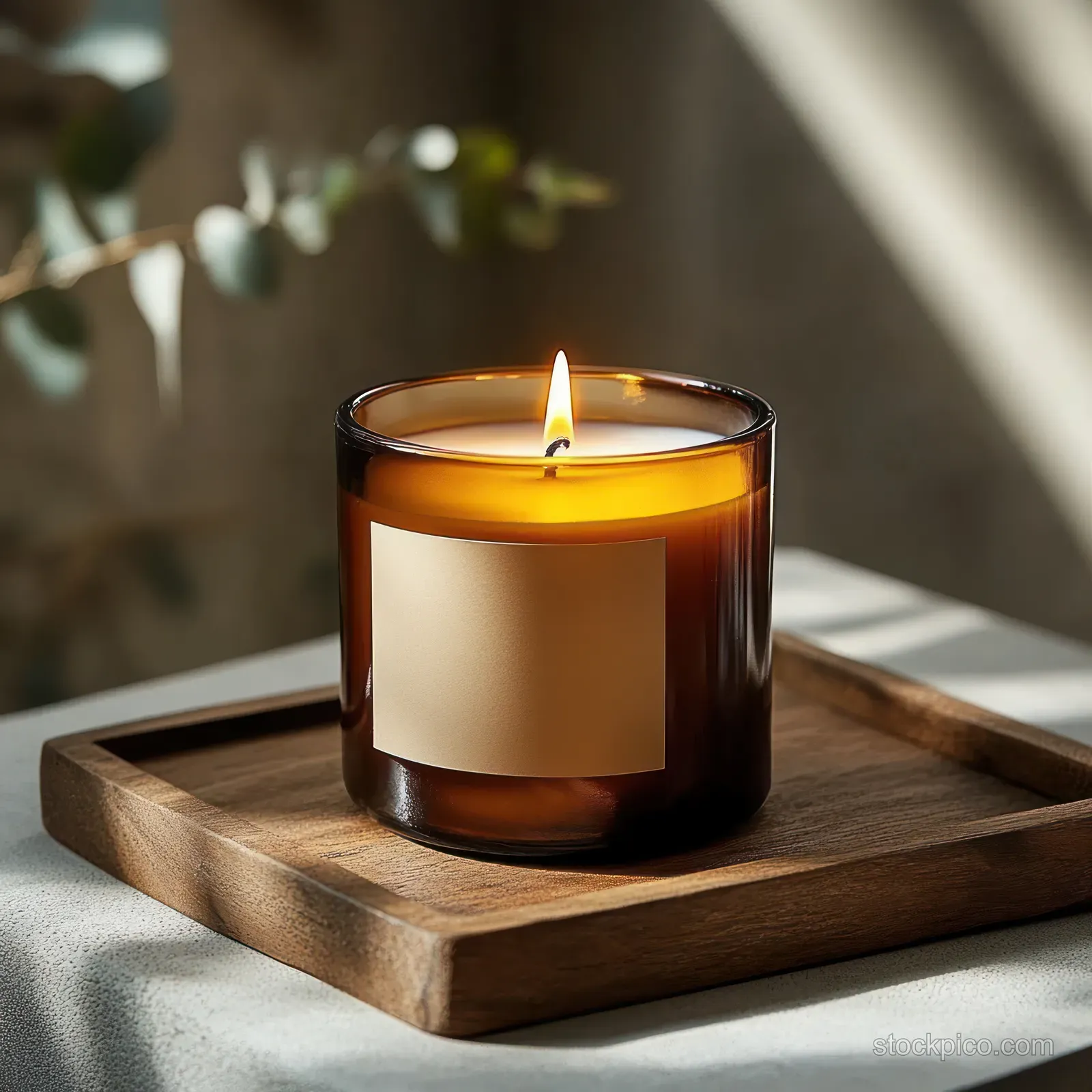 An amber glass candle flickers on a wooden tray with soft daylight and blurred green leaves in the background.