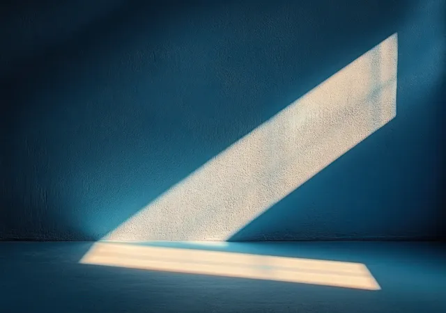 Diagonal sunlight beam casts sharp geometric shadows across a textured blue wall and floor in an empty room.