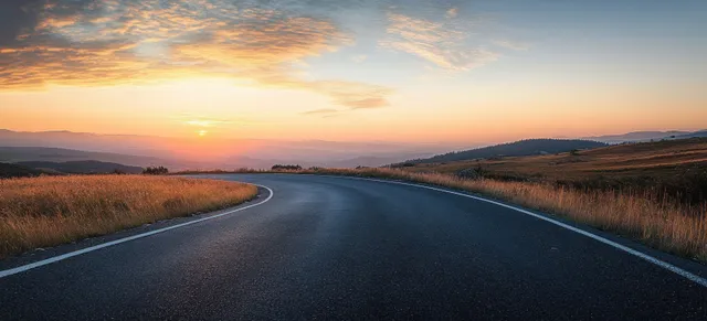 An empty winding mountain road curves through rural hills as the sun sets behind dramatic clouds.