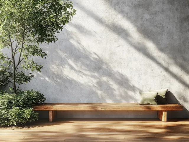 Wooden bench placed along a textured concrete wall with dappled sunlight shadows and green leaves nearby.