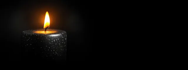 A black candle burns with a flickering flame, casting warm light and deep shadows on a black background.