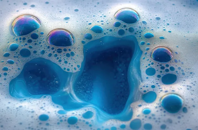Iridescent blue soap bubbles and foam gather on a smooth wet surface with light reflections.