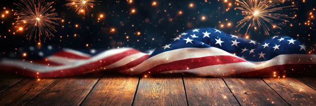 American flag waves on a weathered wooden table while bright fireworks and smoke fill the night sky.