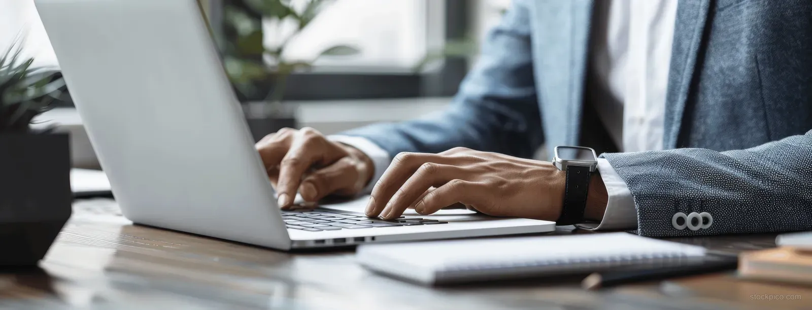 Businessman’s hands typing on a laptop keyboard at a desk beside a notebook in window light.